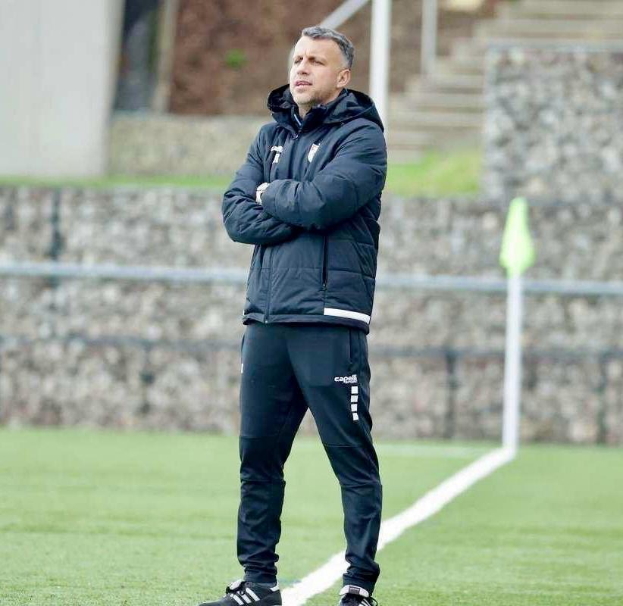 Ein Fußballtrainer auf einem Feld mit verschränkten Armen, der eine schwarze Jacke und Hose trägt, vor einem Zaun, Treppe und Wand während eines Trainings.