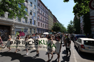 Eine Gruppe von Menschen marschiert auf der Straße in Berlin, hält eine Fahne, mit parkenden Autos auf der rechten Seite, einem Radfahrer auf der linken Seite und Gebäude, Bäumen und einer Schautafel im Hintergrund unter einem sichtbaren Himmel.