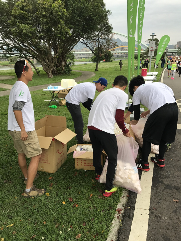 Group of people on a roadside participating in a charity run, surrounded by scattered cardboard boxes, bottles, and other debris, with trees, buildings, and banners in the background under a clear blue sky.