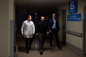 Eine Gruppe verzweifelter Männer geht einen Krankenhausflur entlang, mit einer Tür auf der rechten Seite und Texttafeln auf der linken Seite.