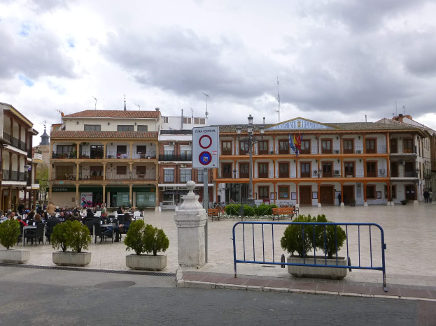 Ein belebter Stadtplatz mit Menschen, die sitzen und stehen, Topfpflanzen, Metallabsperrungen, Straßenlaternen mit Flaggen, ein Schild, Gebäude mit Fenstern und ein bewölkter Himmel.