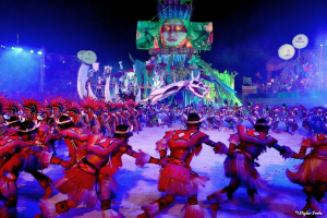 Eine Gruppe von Menschen in bunten Kostümen tanzt auf einer Bühne vor einem sitzenden Publikum, mit Lichtern, Stangen und Luftballons, und ein Bildschirm zeigt eine Person im Hintergrund, während des Karnevals in Rio de Janeiro 2014.