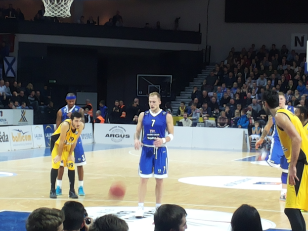Eine Gruppe von Männern in blauen und gelben Uniformen, die Basketball auf einem Court spielen, mit Zuschauern auf den Tribünen und einem Basketballkorb am oberen Ende des Courts.
