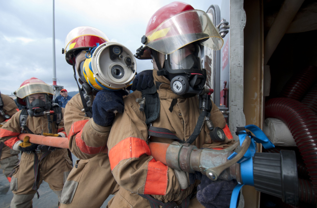 Feuerwehrleute in Schutzausrüstung mit einem, der einen Schlauch hält, eine Stange im Hintergrund und Rohre auf der rechten Seite unter einem bewölkten Himmel.