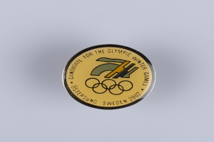 Ein runder Abzeichen mit schwarzem Hintergrund und goldfarbenem Olympia-Logo in der Mitte, mit der Aufschrift "Olympische Winterspiele" in fetter weißer Schrift.