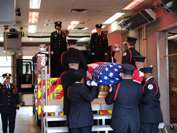 Eine Gruppe uniformierter Männer trägt einen Sarg mit einer amerikanischen Flagge, umgeben von ein paar Männern mit Mützen und einem Löschfahrzeug sowie Deckenlampen im Hintergrund, was auf eine Trauerfeier für einen gefallenen Feuerwehrmann hindeutet.