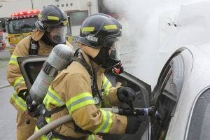 Zwei Feuerwehrleute in Schutzausrüstung verwenden einen Schlauch, um ein brennendes Auto zu löschen, mit mehreren Fahrzeugen und einem Gebäude im Hintergrund, Rauch steigt aus dem Auto.