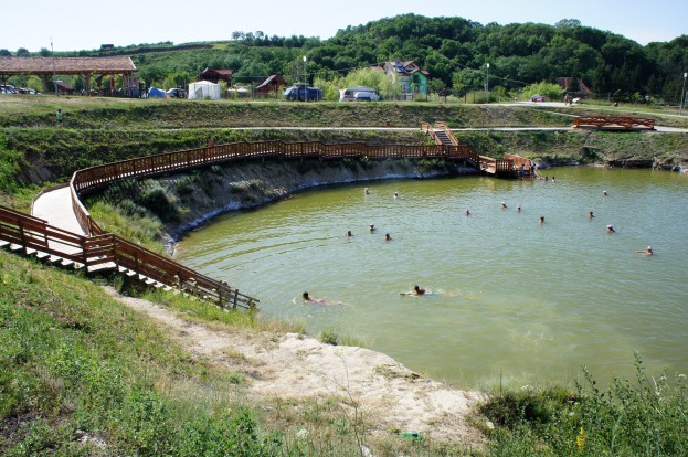 Gruppe von Menschen, die in einem Gewässer schwimmen, mit Brücke, Treppen, Schuppen, Fahrzeugen, Pfählen und üppiger grüner Umgebung unter einem klaren blauen Himmel.
