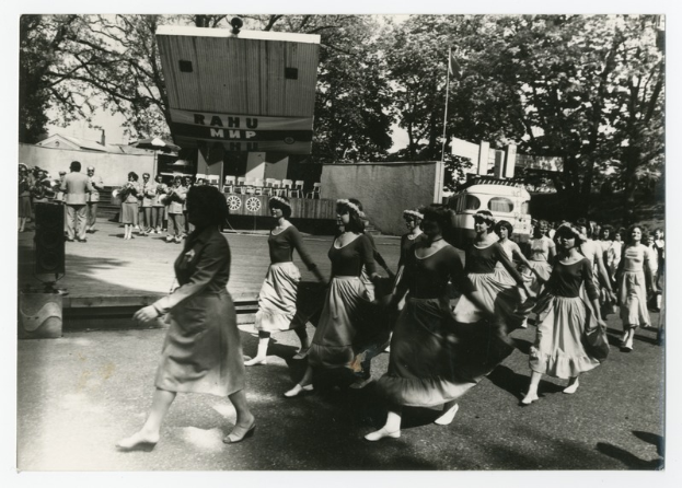 Eine Gruppe von Frauen tanzt auf der Straße, umgeben von einer Menge, einem Lautsprecher, einem Fahrzeug, einer Tafel mit Text, einem Fahnenmast, Gebäuden und einem bewölkten Himmel.
