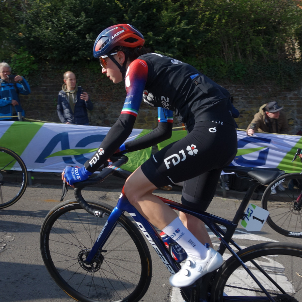 Professionelle Radrennfahrerin Katie Archibald fährt ein Fahrrad auf einer Straße, trägt einen Helm, eine Schutzbrille, Handschuhe und Schuhe und hat eine Menschenmenge neben sich.