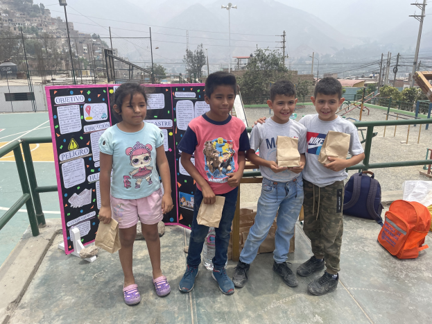 Gruppe von Kindern auf einem Basketballfeld, die Papiertütten halten, mit Gegenständen auf der rechten Seite und einer Tafel mit Text auf der linken Seite, vor einem Hintergrund aus Zäunen, Masten, Straßenlampen, elektrischen Masten mit Drähten, Bäumen, Gebäuden, Hüllen und Himmel.