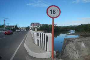 Ein Tempolimit-Schild am Rande einer Straße neben einem Fluss, mit Fahrzeugen, einer Barriere, Bäumen, Gebäuden, Strommästen mit Drähten und einem bewölktem Himmel im Hintergrund.