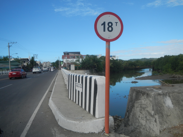 Ein Tempolimit-Schild am Rande einer Straße neben einem Fluss, mit Fahrzeugen, einer Barriere, Bäumen, Gebäuden, Strommästen mit Drähten und einem bewölktem Himmel im Hintergrund.