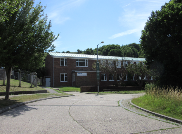 Ein Schulgebäude mit Fenstern, einem Straßenschild, einem Schild, einer Baumgruppe, Gras, einem Zaun und einem bewölkten Himmel, das in der Mitte eines Parks umgeben von saftigem Gras steht.