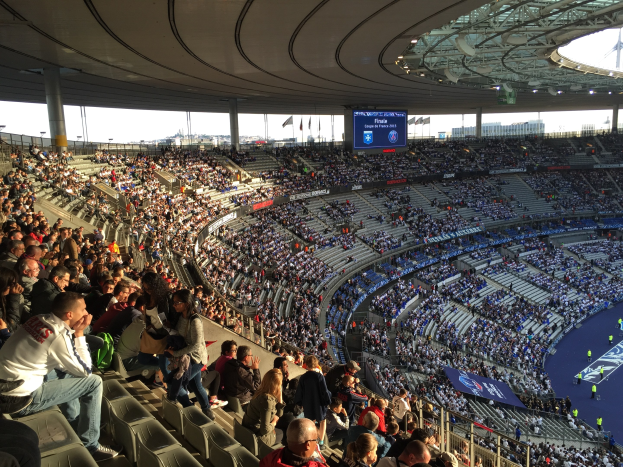 Gro├če Zuschauermenge in der Allianz Arena bei einem Fuballspiel, mit einer Bühne auf der rechten Seite, Fahnen, Masten, einem Bildschirm und sichtbarem Himmel im Hintergrund.