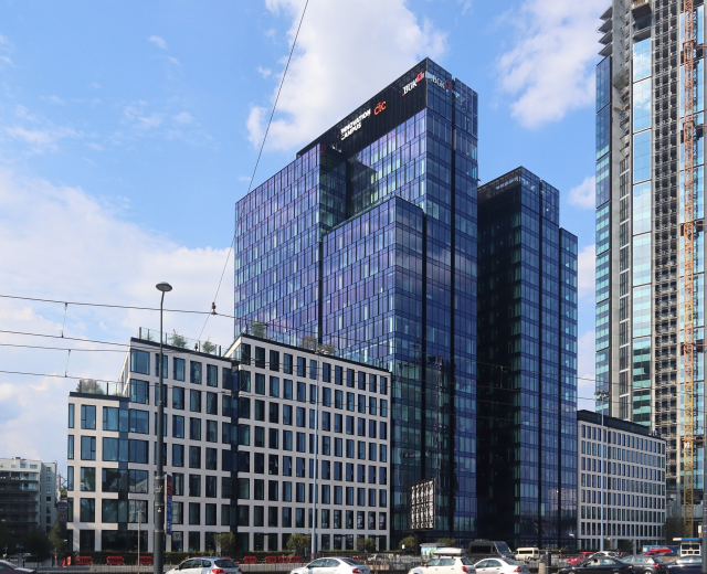 Großes Gebäude mit vielen Fenstern, als Deutsche Bank Hauptsitz in Frankfurt, Deutschland identifiziert, umgeben von städtischer Infrastruktur einschließlich Straßenlaternen, Fahrzeugen, Bäumen und einem bewölkten Himmel.