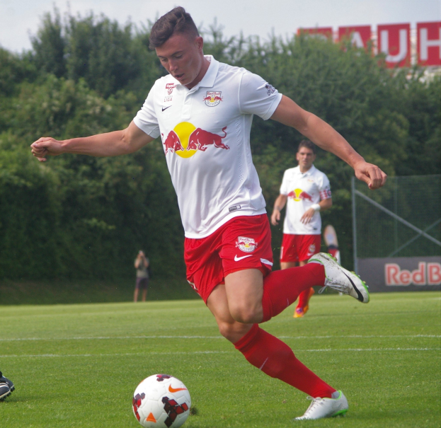 Ein professioneller Füßballspieler in roter und weißer Uniform tritt einen Füßball auf einem grünen Feld mit Bäumen und einem klaren blauen Himmel im Hintergrund, mit einem "RB Leipzig"-Schild im Bild.