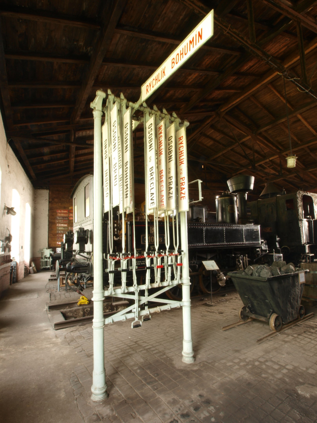Ein Bahnsteig mit einem Zug auf den Gleisen, einem Rollstuhl auf dem Boden, einer Signalbox in der Mitte, einem Pfahl mit einer daran befestigten Tafel, einer Wand auf der linken Seite und einer Decke darüber.