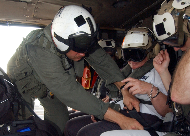 Eine Gruppe von Männern in militärischer Uniform und Helmen sitzt im Heck eines Hubschraubers, wobei einer der Männer eine Brille und eine Uhr trägt und andere Taschen tragen, neben einem Verbandkasten.