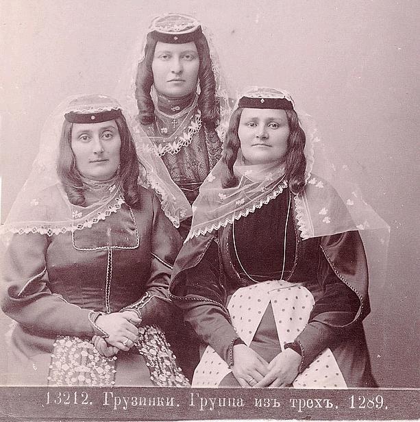 Drei Frauen in traditionellen russischen Hochzeitskleidern mit aufwändiger Stickerei und Perlen posieren vor einer weißen Wand in einem Schwarz-Weiß-Foto.