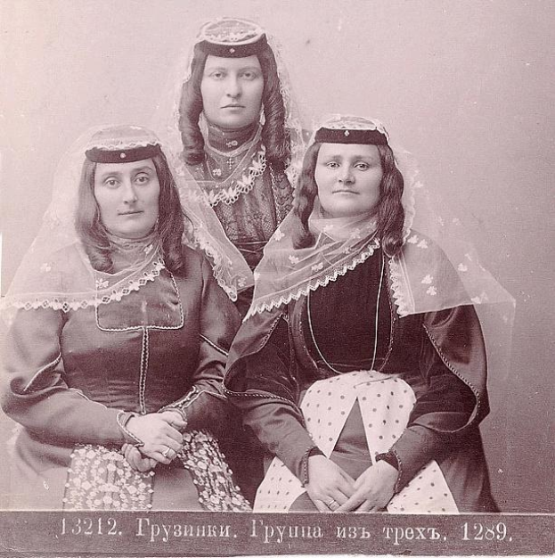 Drei Frauen in traditionellen russischen Hochzeitskleidern mit aufwändiger Stickerei und Perlen posieren vor einer weißen Wand in einem Schwarz-Weiß-Foto.
