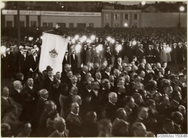 Schwarzes und weißes Foto einer großen Menschenmenge vor einem Gebäude, mit einer Person, die eine Fahne hält, Pfosten und Lichter im Hintergrund und Text unten mit der Aufschrift "Gustav Klimts Rede an der Berliner Mauer".