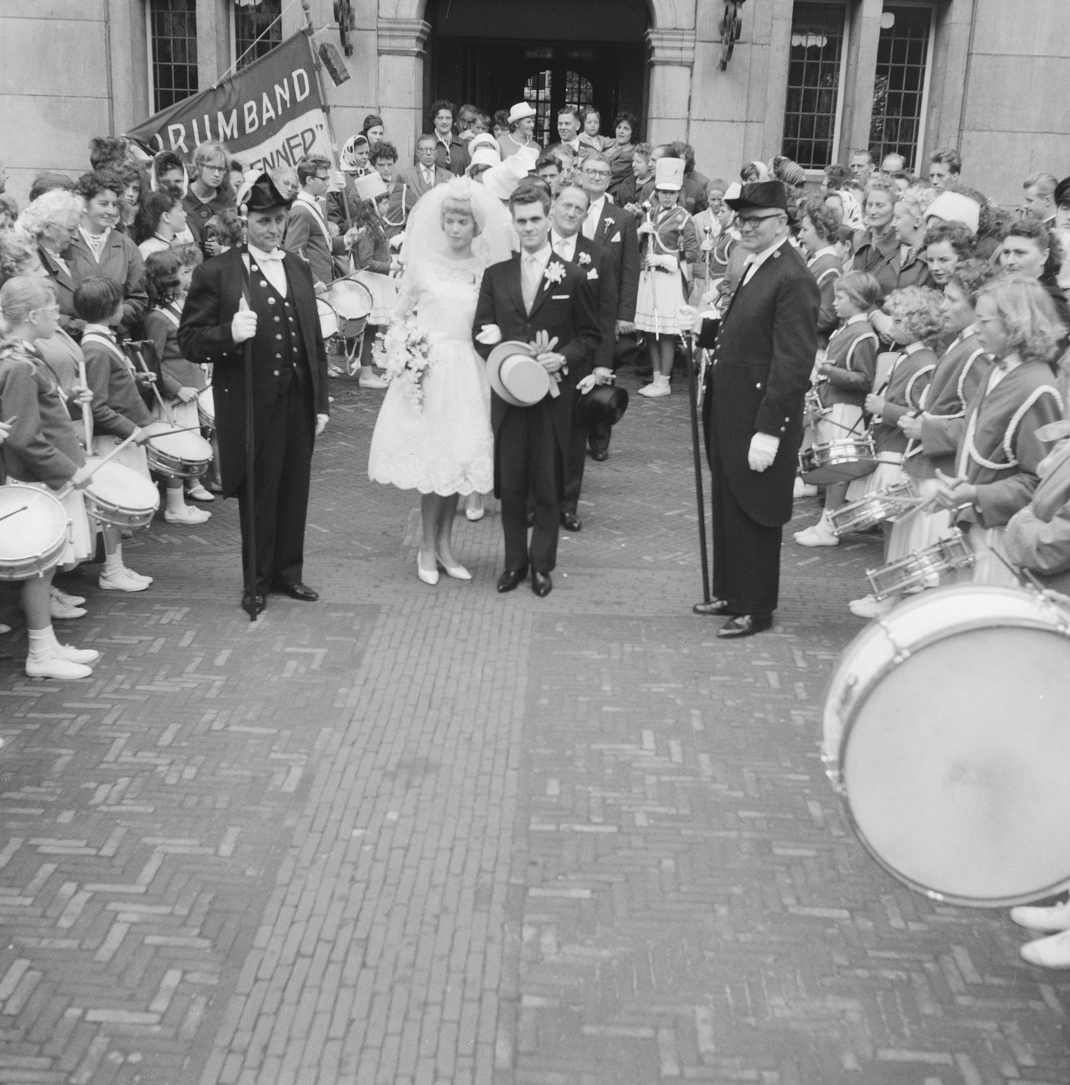 Schwarzes und weißes Foto einer Braut in einem weißem Kleid und Schleier und Bräutigam mit einem Strauß Blumen, der eine Straße öberquert, vorbei an einer Menge, einige spielen Instrumente, mit einem Banner und einem Gebäude im Hintergrund.
