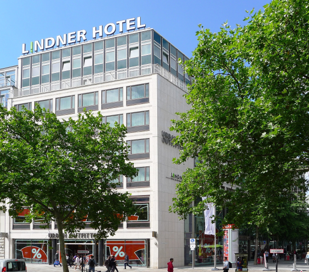Großes Glasfrontgebäude mit der Aufschrift "Lindner Hotel" in Berlin, mit Bäumen, Fahrzeugen, Fußgängern und Himmel im Hintergrund.