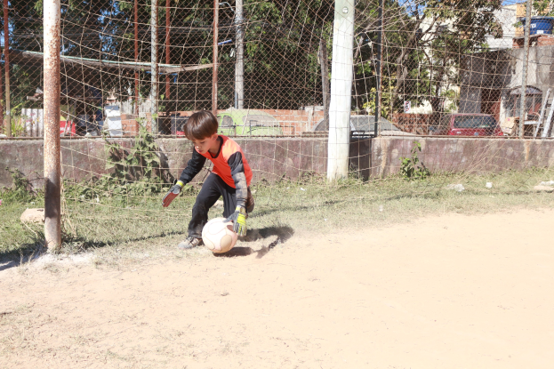 Ein junger Junge spielt Füßball auf einem Schmutzfeld mit einem bewölktem Himmel, umgeben von Gras, Pflanzen, Polen, einem Zaun, einer Wand, Bäumen, Fahrzeugen und Gebäuden.