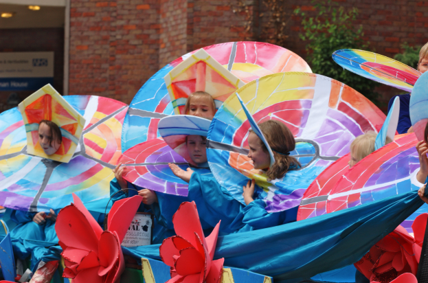 Eine Gruppe von Kindern in bunten Kostümen fährt auf einem Paradewagen, hält Schirme, mit einem Gebäude, Pflanzen und Lichtern im Hintergrund.