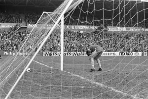 Ein Schwarz-Weiß-Foto eines Fötballspielers, der vor einem Publikum in einem Stadion einen gehalten hat, mit einem Tor und einem Ball in der Nähe und Bannern mit Text im Hintergrund.