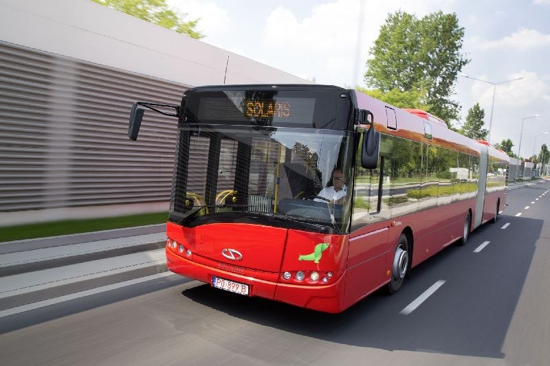 Roter Bus fährt auf einer Straße neben einem Gebäude, mit einer Person im Inneren; eine Wand auf der linken Seite, Bäume, Laternenpfähle und ein bewölkter Himmel im Hintergrund.