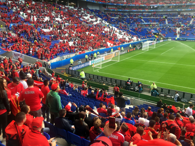 Eine große Menschenmenge in einem Stadion schaut bei einem Fußballspiel zu, mit Sitzplätzen und Stehplätzen, Fahnen, einem Tor und Deckenleuchten.