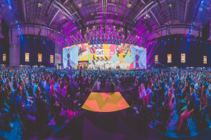 Eine große Menschenmenge, die auf Stühlen vor einer hell erleuchteten Bühne sitzt, mit mehreren stehenden Individuen, wahrscheinlich auf einem Online-Marketing-Rockstars-Event.