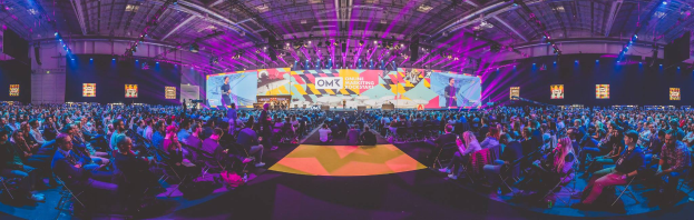 Eine große Menschenmenge, die auf Stühlen vor einer hell erleuchteten Bühne sitzt, mit mehreren stehenden Individuen, wahrscheinlich auf einem Online-Marketing-Rockstars-Event.