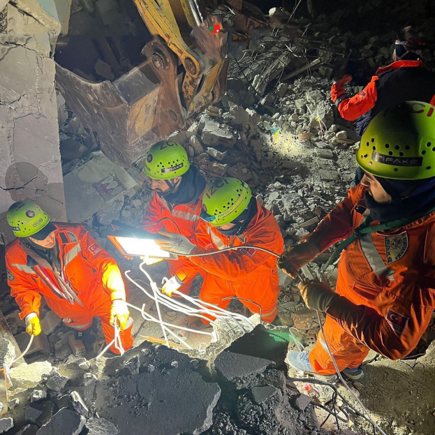 Menschen in orangefarbener Uniform und grünem Helm bei der Arbeit an Schutt mit einem Kran im Hintergrund bei einem Einsatz nach dem Einsturz eines Gebäudes.
