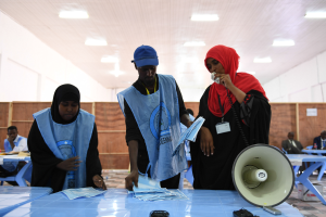 Eine Gruppe von Menschen, die um einen Tisch mit einem Megaphon, Papieren und anderen Gegenständen stehen, mit einigen Menschen im Hintergrund sitzend und stehend, und einer Decke mit Lichtern an einer Wahllokal.