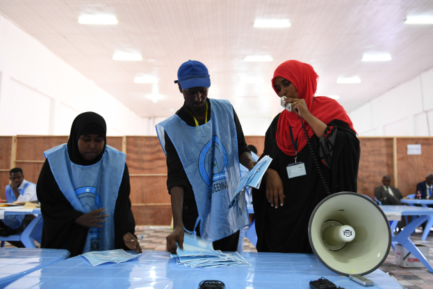 Eine Gruppe von Menschen, die um einen Tisch mit einem Megaphon, Papieren und anderen Gegenständen stehen, mit einigen Menschen im Hintergrund sitzend und stehend, und einer Decke mit Lichtern an einer Wahllokal.