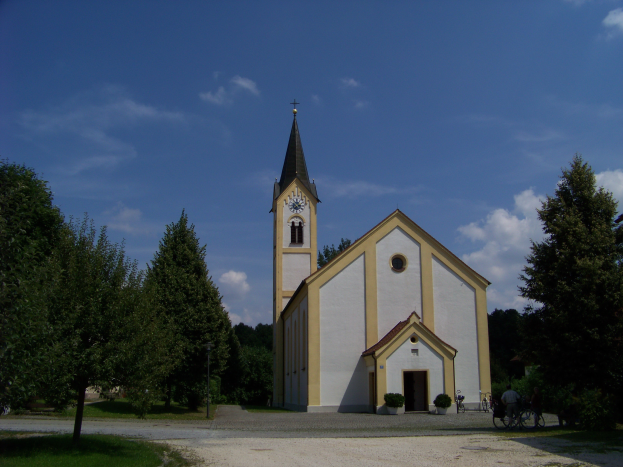 Eine Kirche mit Turm und Uhr steht in einem Feld umgeben von Häusern, Töpfen mit Pflanzen, Bäumen, Gras, einem Weg, Radfahrern und unter einem bewölkten Himmel.