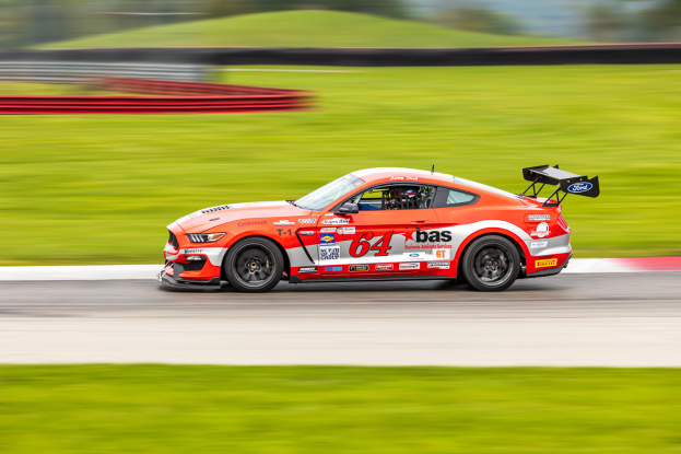 Ein Ford Mustang GT Rennauto mit Text und Zahlen darauf fährt auf einer Rennstrecke, mit einem Helmträger im Sitz, der Hintergrund ist unscharf, um Geschwindigkeit und Bewegung zu zeigen.